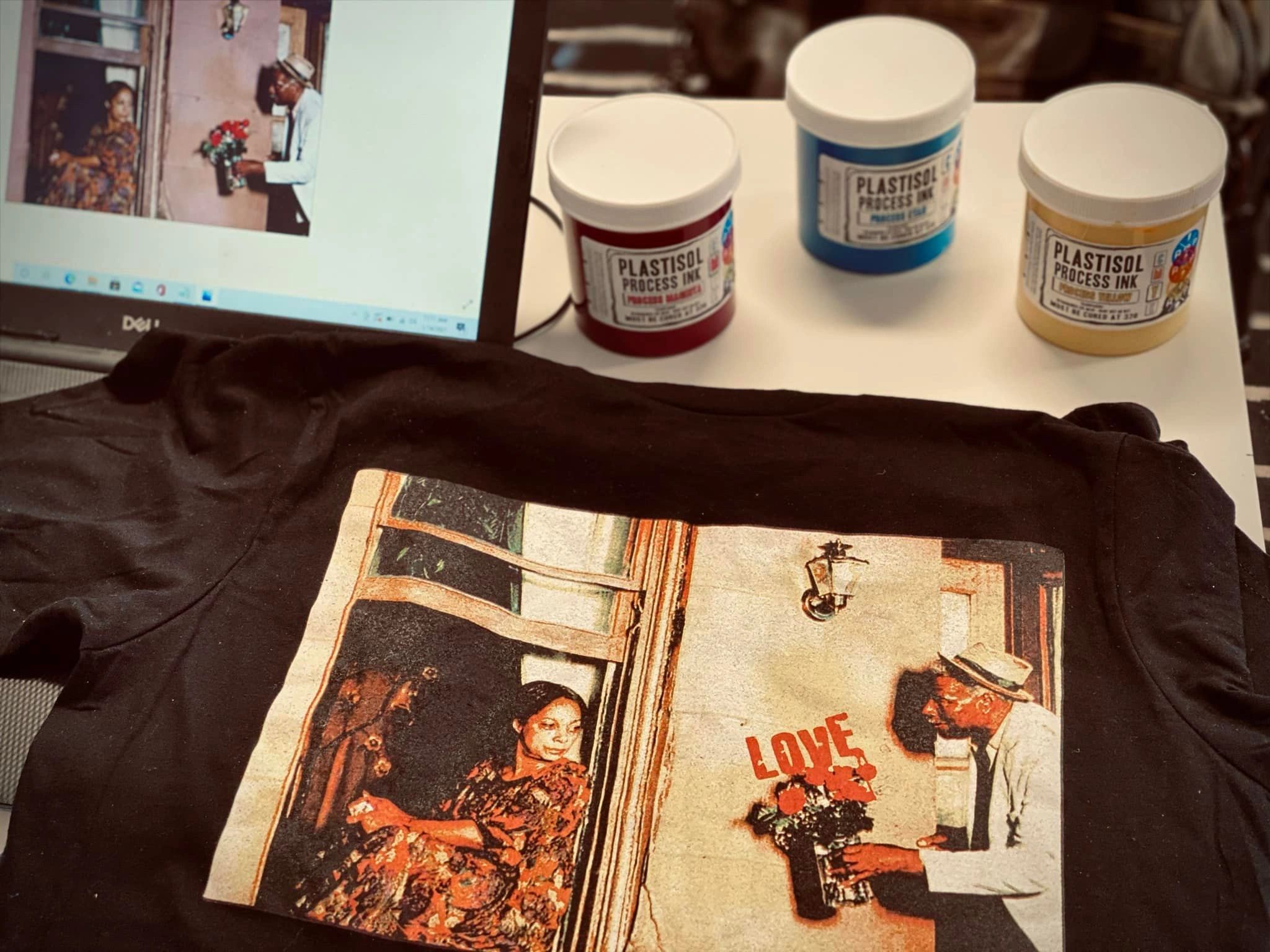 A worktable view brings together printed apparel, color reference artwork and ink containers, offering a glimpse of design and production materials.