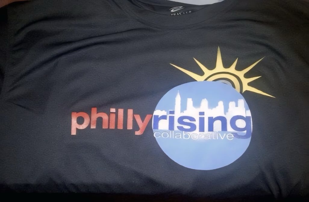 A close-up of the Philly Rising graphic on dark fabric, with a bright yellow sun and contrasting blue and red lettering.