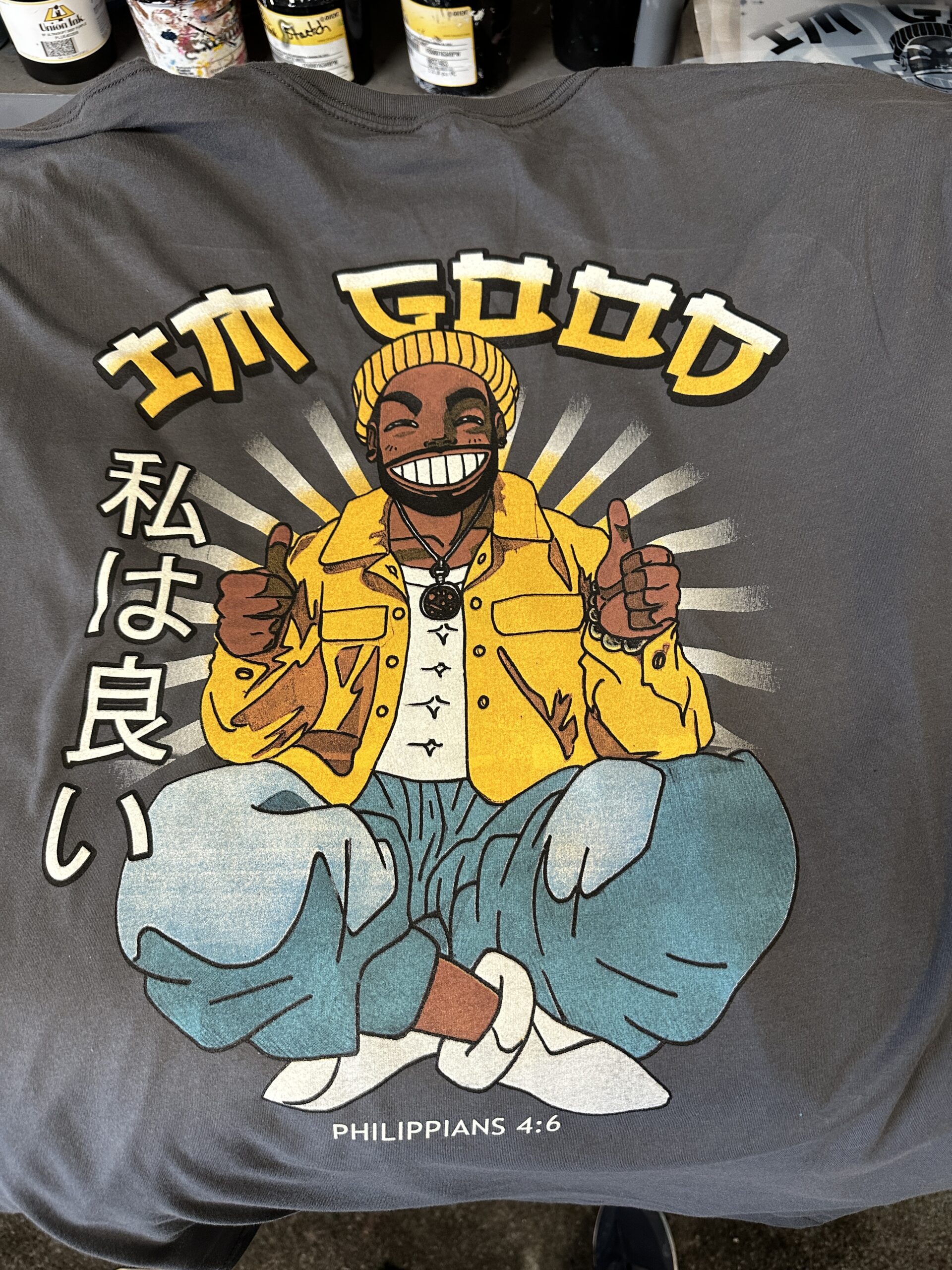 A dark tee combines gold “I’m Good” lettering with an illustrated seated figure and vertical characters at the side.